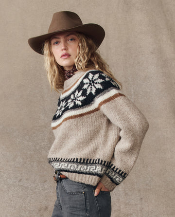 A person wears The Snowflake Fairisle Pullover in Oat from THE GREAT. H25 COLLECTION, paired with a wide-brimmed brown hat and grey jeans, standing against a neutral background.