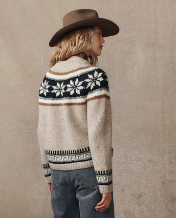 A person wears THE GREAT. H25 COLLECTION's The Snowflake Fairisle Pullover in Oat with a brown hat and blue jeans, standing against a neutral background.