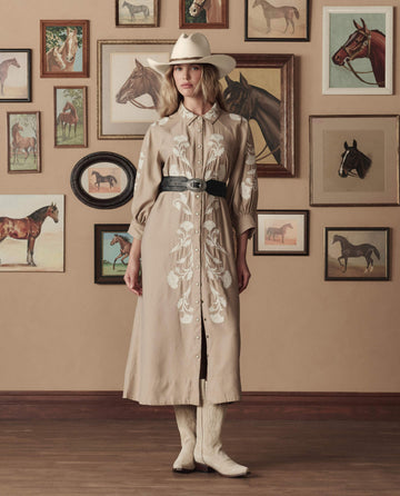 A woman wears THE GREAT. STETSON SP26’s Western Embroidered Herd Dress in soft tan with cream embroidery, a white cowboy hat, and boots, standing before a wall of framed horse paintings and photos.