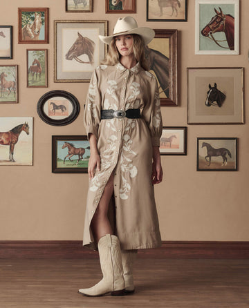 A woman wears THE GREAT. STETSON SP26's The Western Embroidered Herd Dress in soft tan with cream embroidery, standing indoors by a wall of framed horse art, accessorized with a cowboy hat and boots.