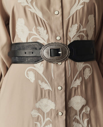 Close-up of a beige shirt with white floral embroidery, styled with THE GREAT. STETSON SP26 "The Tooled Waist Belt" in black with a decorative silver buckle at the waist.