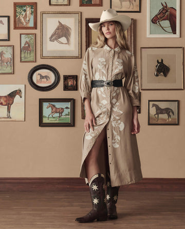 A woman in a beige embroidered shirt dress, cowboy hat, and THE GREAT. STETSON SP26 The Cowboy Boot (Leather Floral) - Brown Floral stands before a wall adorned with horse-themed art.