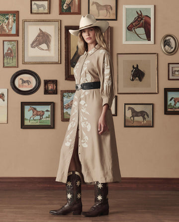 A woman in a beige embroidered dress, cowboy hat, and THE GREAT. STETSON SP26 The Cowboy Boot (Leather Floral) in Brown Floral stands before a wall of framed horse paintings.