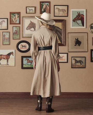 Wearing THE GREAT. STETSON SP26's Western Embroidered Herd Dress in soft tan with cream embroidery, a person stands facing a wall covered in framed horse paintings and photos.