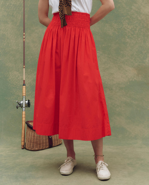 A person stands holding a fishing rod, wearing THE GREAT. SU25’s The Viola Skirt in Hot Red, paired with white sneakers and an anklet, beside a woven basket—effortlessly highlighting women's fashion against a neutral backdrop.