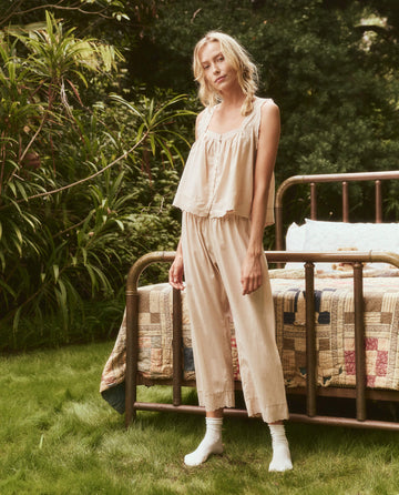 A woman in light-colored loungewear, including The Eyelet Tank in Soft Rosette by THE GREAT. SP24 SLEEP, stands near a metal-framed bed with a quilt in an outdoor setting surrounded by greenery.