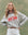 A person with long blonde hair wears THE GREAT. SU25 The Teammate Sweatshirt in Soft Heather Grey with a Smooth Sailin graphic, paired with white pants and a brown belt, standing against a green backdrop.