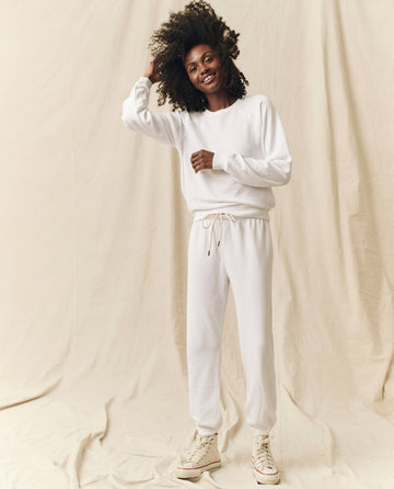 A person with curly hair smiles while standing. They are wearing The Shrunken Sweatshirt in True White from THE GREAT. FALL 24 KNITS, a white sweatshirt and sweatpants set with white sneakers, and the background is a plain light-colored fabric.