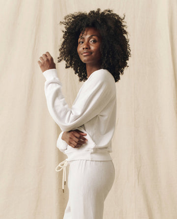 A person stands against a light-colored backdrop, wearing The Shrunken Sweatshirt in True White from THE GREAT. FALL 24 KNITS, paired with matching pants. With curly hair and a neutral expression, they look slightly to the side, evoking the timeless charm of vintage sweatshirts.