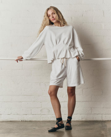 A woman in THE GREAT. SP26 DANCER BODIES The Relaxed Sport Short in True White and a matching top stands by a white brick wall, wearing black ballet flats with crisscross straps.