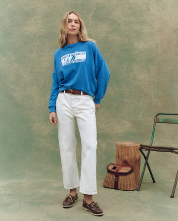 A woman stands against a textured backdrop, wearing THE GREAT. SP25 D1 The Wayne Jean in True White. A wicker bag, wooden stump, and folding chair are placed beside her.