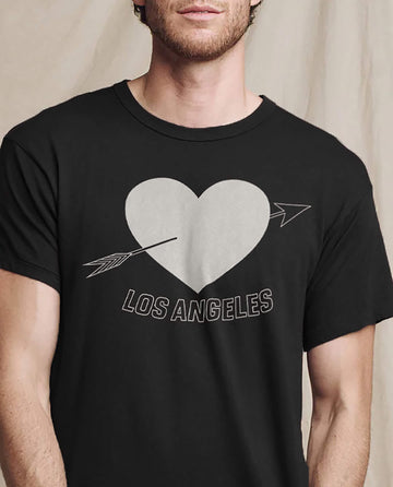 Wearing THE GREAT. SP25 I LOVE LA's Men's Boxy Crew, a T-shirt in Almost Black featuring an LA heart graphic with a playful arrow and "Los Angeles" text, this design captures a relaxed vibe.