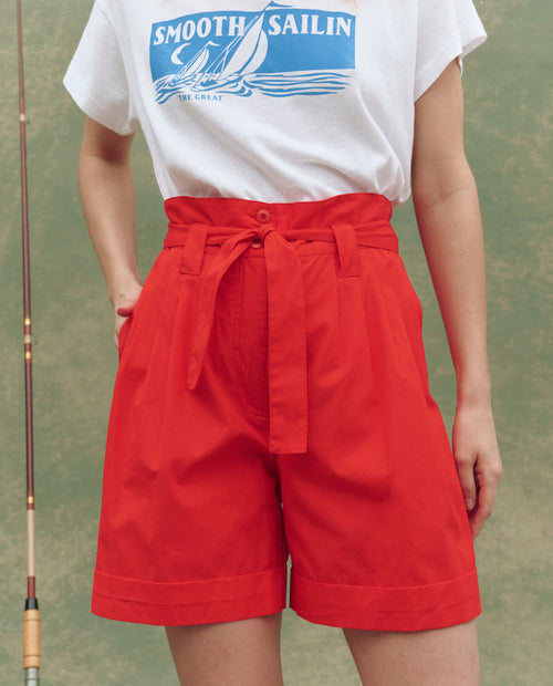 Person wearing a white "Smooth Sailin" graphic tee and THE GREAT. SU25 The Pleated Trouser Short in Hot Red, featuring a high-waisted fit, pleats, and a tie belt, standing with one hand in their pocket.