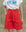 Person wearing a white "Smooth Sailin" graphic tee and THE GREAT. SU25 The Pleated Trouser Short in Hot Red, featuring a high-waisted fit, pleats, and a tie belt, standing with one hand in their pocket.