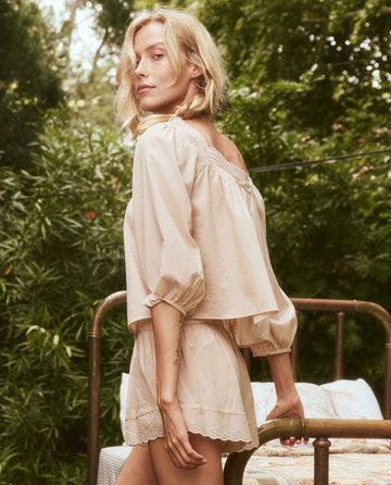 A woman with blonde hair is wearing The Eyelet Button Sleep Top in Soft Rosette by THE GREAT. SP24 SLEEP, paired with matching shorts, standing outdoors against a backdrop of greenery and a metal bed frame.
