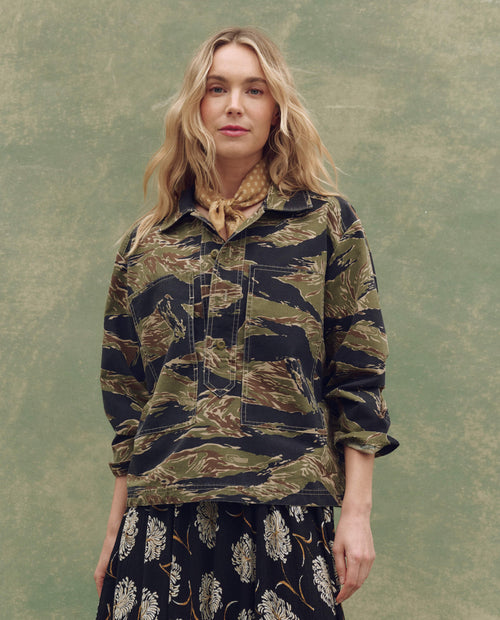 A woman with wavy blonde hair stands before a green backdrop, styled in THE GREAT. SU25's The Railway Shirt—Camo Surplus—as a jacket, with a patterned scarf and a black floral skirt.