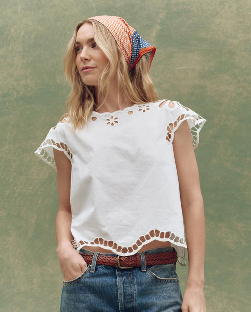A woman in THE GREAT. SU25 The Super Bloom Eyelet Top in white with blue jeans and a red patterned headscarf stands before a green textured backdrop, embodying a vibrant super bloom-inspired style.
