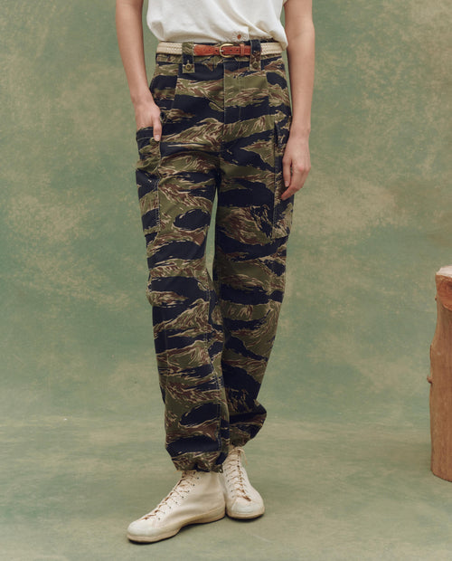 A person stands on a green background wearing a white shirt, THE GREAT. SU25 The Convoy Pant - Camo Surplus, styled with a brown belt and white high-top sneakers.