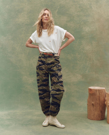 A woman stands before a green backdrop, wearing a white t-shirt, THE GREAT. SU25 The Convoy Pant in Camo Surplus, white sneakers, a brown belt, and with wooden stumps to her right.