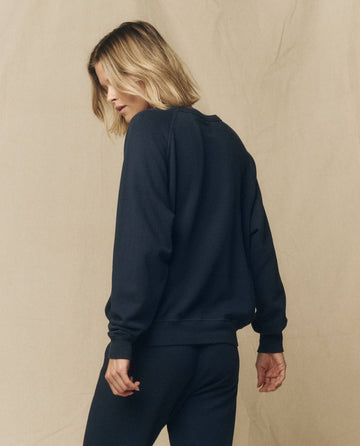 A person with shoulder-length blonde hair is standing with their back facing the camera, wearing "The College Sweatshirt" in True Navy by THE GREAT. FALL 23 KNITS and dark pants, set against a plain beige backdrop.