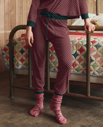 A person stands beside a metal bed, wearing THE GREAT. H25 SLEEP’s The Jersey Long John in the Daisy Tie Print; a quilted bedspread appears in the background.