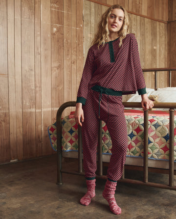 A woman in THE GREAT. H25 SLEEP The Crop Sleep Henley in Daisy Tie Print and socks stands by a bed with a patchwork quilt in a wood-paneled room.