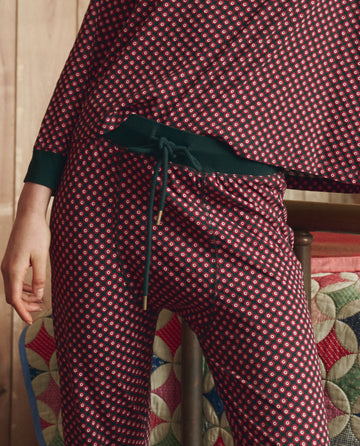 Someone wearing THE GREAT. H25 SLEEP’s The Jersey Long John in Daisy Tie Print stands beside a quilted blanket draped over a wooden chair.