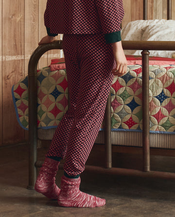 Wearing THE GREAT. H25 SLEEP's The Jersey Long John in Daisy Tie Print and socks, a person stands beside a metal-framed bed with a colorful quilt in a wooden room.