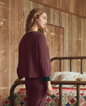 A person with wavy blonde hair stands by a metal bed in a wood-paneled room, wearing matching patterned pajamas and THE GREAT. H25 SLEEP’s The Crop Sleep Henley in Daisy Tie Print.