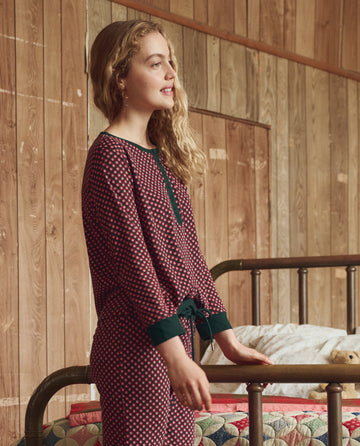 A woman wears The Crop Sleep Henley in Daisy Tie Print by THE GREAT. H25 SLEEP, standing beside a metal bed with a quilted blanket in a cozy, wood-paneled room.