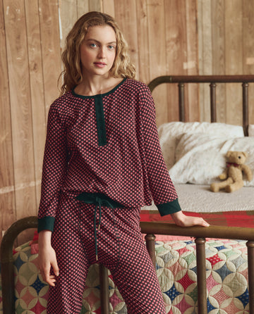 A woman in THE GREAT. H25 SLEEP The Crop Sleep Henley -- Daisy Tie Print stands by a metal-frame bed decorated with a quilt and teddy bear, set against a wooden wall background.