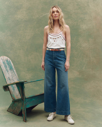 A woman stands by a weathered wooden chair against a green mottled background, wearing THE GREAT. SU25's The Beaded Slim Tank in Washed White, wide-leg blue jeans, a brown belt, and white sneakers.
