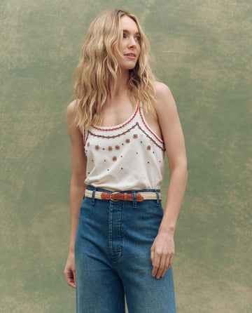 A woman with wavy blonde hair wears THE GREAT. SU25 The Beaded Slim Tank in Washed White and high-waisted blue jeans, standing against a plain textured green background.