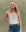 A woman stands against a mottled green background, wearing THE GREAT. SU25 The Beaded Slim Tank in Washed White, styled with blue high-waisted jeans and a woven belt.