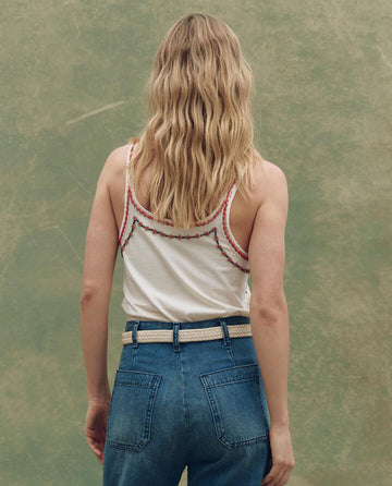 A woman with wavy blonde hair faces away, wearing THE GREAT. SU25 The Beaded Slim Tank in Washed White with red trim and high-waisted blue jeans against a greenish background.