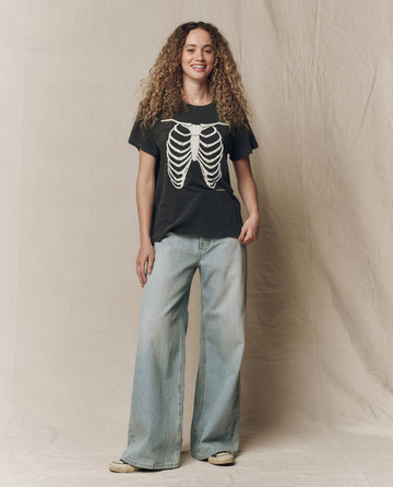 A person with long curly hair wears THE GREAT. F26 HALLOWEEN GRAPHICS “The Boxy Crew w/ Skeleton Graphic” in washed black, paired with loose light blue jeans, and stands against a neutral fabric backdrop.