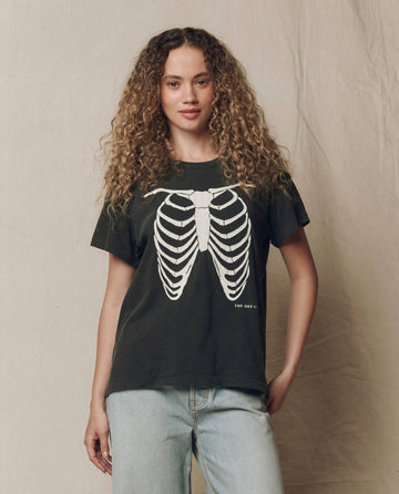 A woman with long curly hair wears THE GREAT.’s The Boxy Crew w/ Skeleton Graphic in washed black, featuring a bold white ribcage graphic on the front, paired with light blue jeans and standing against a neutral fabric background.
