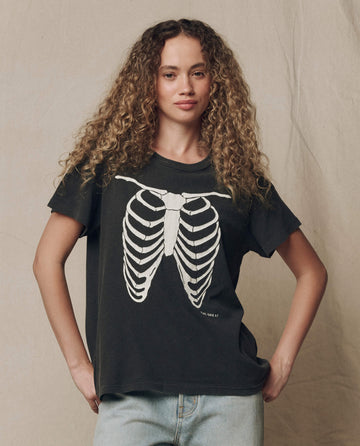 An individual with long curly hair is seen standing against a neutral fabric background, wearing THE GREAT. F26 HALLOWEEN GRAPHICS’ “The Boxy Crew w/ Skeleton Graphic” t-shirt in washed black.