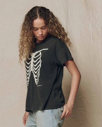 A person with curly hair wears THE GREAT.’s The Boxy Crew w/ Skeleton Graphic in Washed Black from the F26 HALLOWEEN GRAPHICS collection, paired with light blue jeans, standing against a plain beige backdrop.