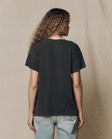 A person with long curly hair, wearing THE GREAT. F26 HALLOWEEN GRAPHICS The Boxy Crew w/ Skeleton Graphic in washed black and light blue jeans, stands with their back to the camera against a beige fabric backdrop.