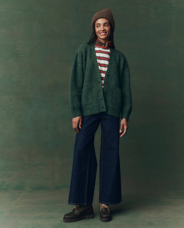 A person stands against a green background, smiling and looking to the side while wearing THE GREAT. F25 D2 The Brushed Melange Cardigan in Meadow Green, a striped shirt, brown beanie, wide-leg jeans, scarf, and chunky shoes.