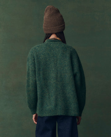 A person stands facing away from the camera, wearing THE GREAT. F25 D2's The Brushed Melange Cardigan in Meadow Green with dark pants against a greenish background.