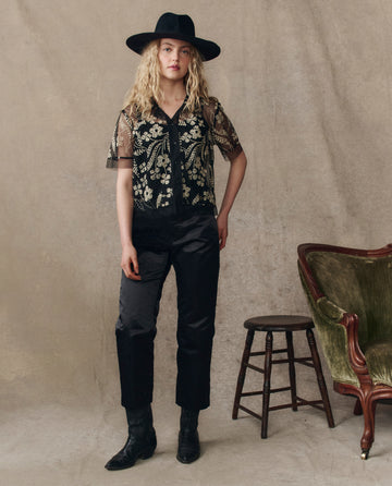 A woman wears THE GREAT. H25 COLLECTION’s The Cotillion Top in Brocade Floral, paired with black pants, boots, and a black hat, standing by a wooden stool and green armchair against a neutral backdrop.