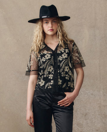A person wears THE GREAT. H25 COLLECTION’s Cotillion Top in Brocade Floral, paired with black pants and a wide-brim hat, standing against a plain beige background.