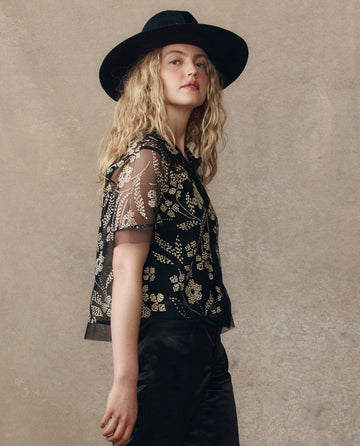 A person with long, wavy hair wears a wide-brimmed black hat, black pants, and THE GREAT. H25 COLLECTION's The Cotillion Top in Brocade Floral, standing against a plain textured background.