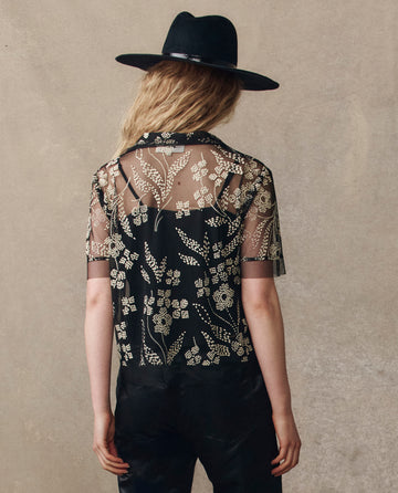 A person with wavy blonde hair, back to camera, wears THE GREAT. H25 COLLECTION's The Cotillion Top in Brocade Floral—sheer black with gold floral embroidery—layered over a black blouse and paired with a black hat.