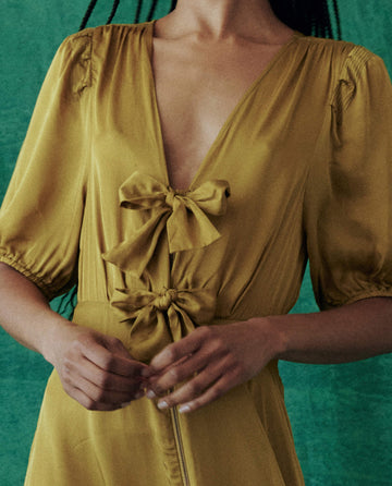 A person wearing The Gilded Dress - Gold Leaf from THE GREAT. HOL 24 stands elegantly in front of a green background, embodying the essence of luxury fashion.