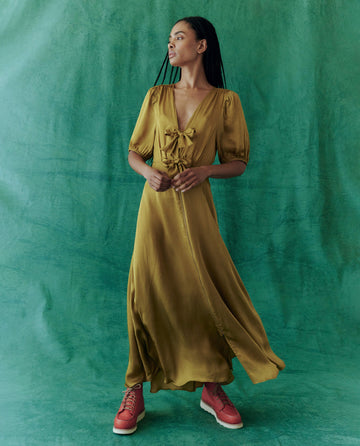 Standing against a green backdrop, an individual embodies the essence of luxury fashion in The Gilded Dress by THE GREAT. HOL 24—Gold Leaf—and red shoes, gazing to the left with an air of elegance.