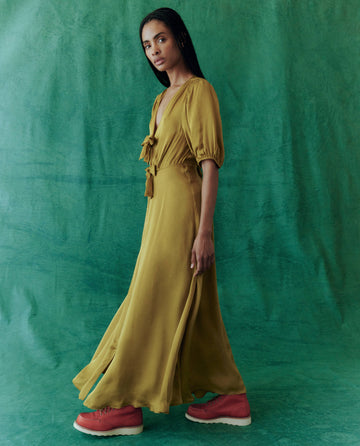 A person dressed in *The Gilded Dress - Gold Leaf* from THE GREAT. HOL 24, accompanied by red shoes, stands against a green background.