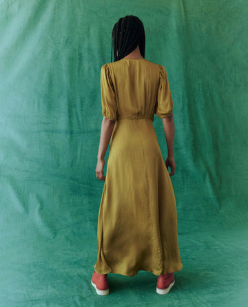 A person in a luxurious, full-length Gold Leaf dress from THE GREAT. HOL 24 stands against a green backdrop, showcasing elegant evening wear complemented by eye-catching pink shoes.